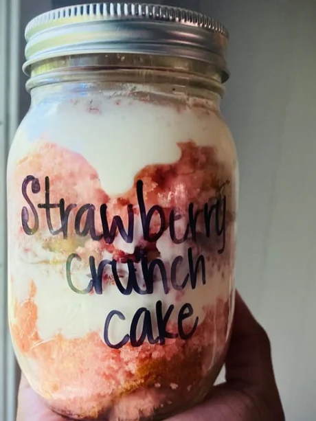 A Strawberry Crunch cake jar held up in one hand, pink cake and cream layered in a mason jar